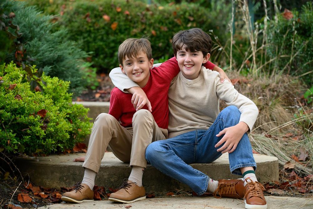 Two boys sit close together on outdoor stone steps surrounded by greenery in a garden setting.
