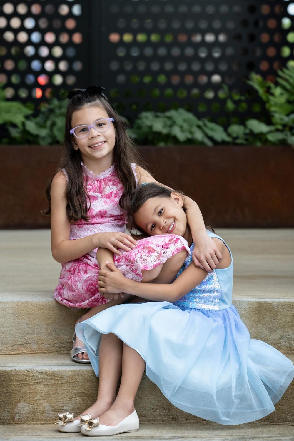 Two children in dresses sit together on concrete steps with one leaning affectionately on the other.