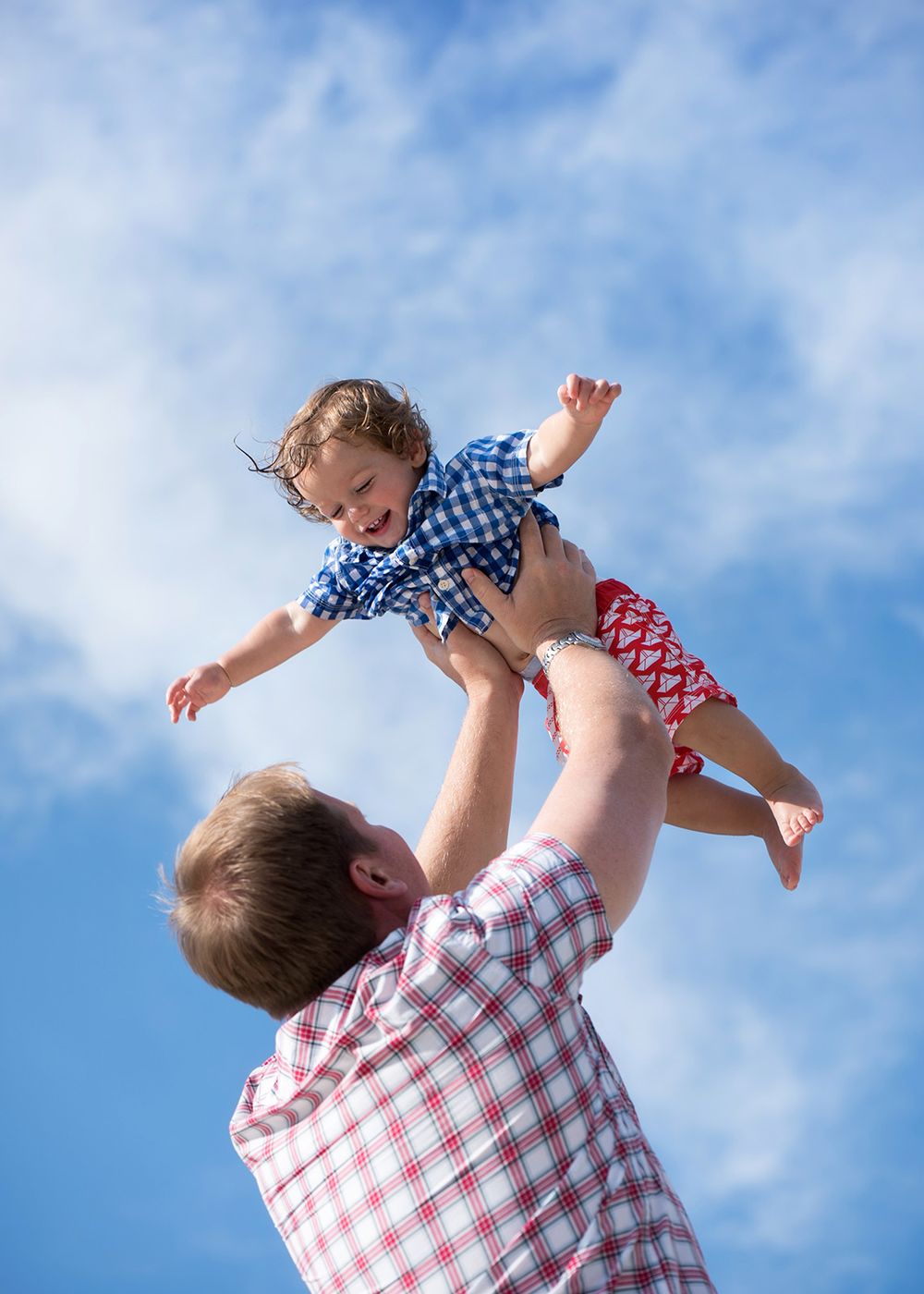 A parent lifts a small child into the air against a bright blue sky with puffy white clouds.