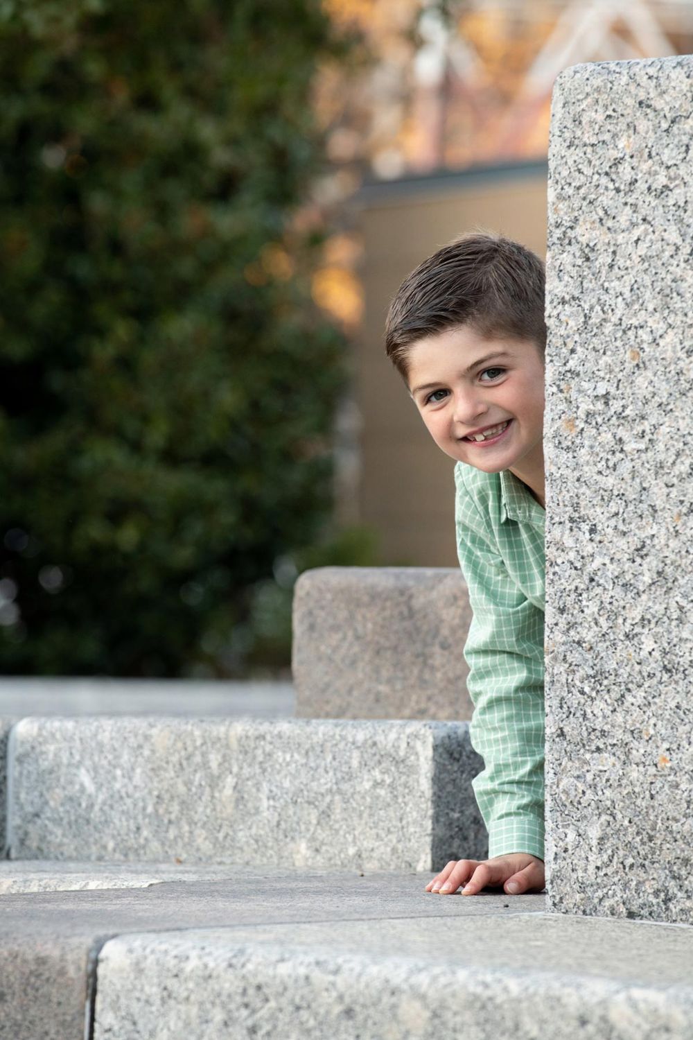 A person in a mint green shirt peeks playfully from behind a concrete wall in an outdoor setting.