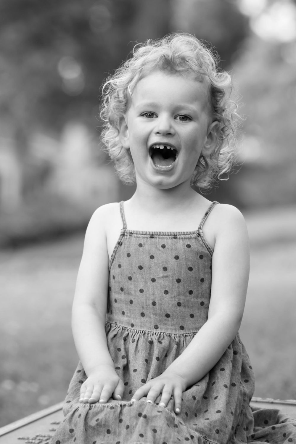 A lively toddler in a polka dot dress laughs and plays outdoors in a black and white photograph.