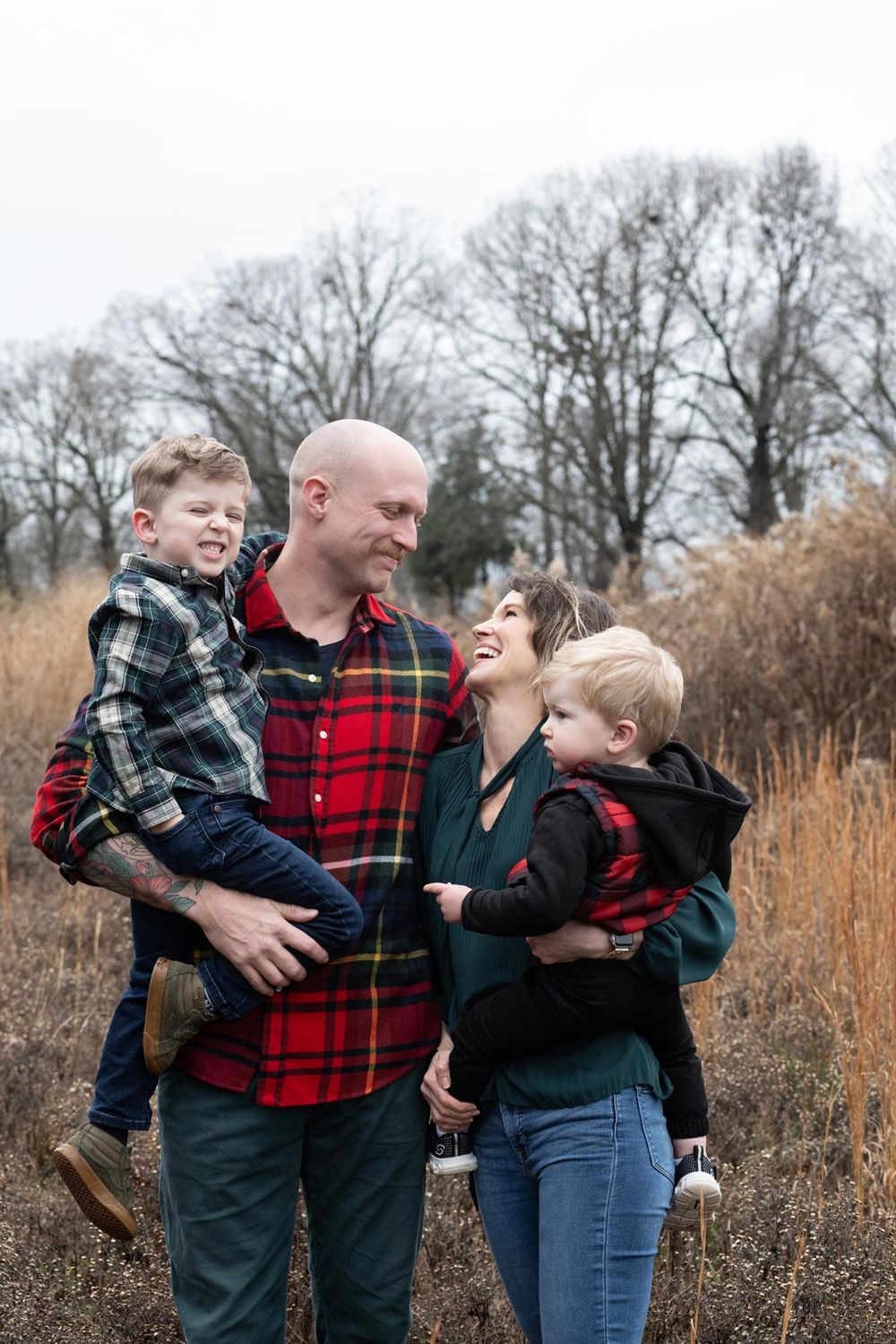 Family wearing plaid and winter clothing poses together in tall autumn grass with bare trees in background.