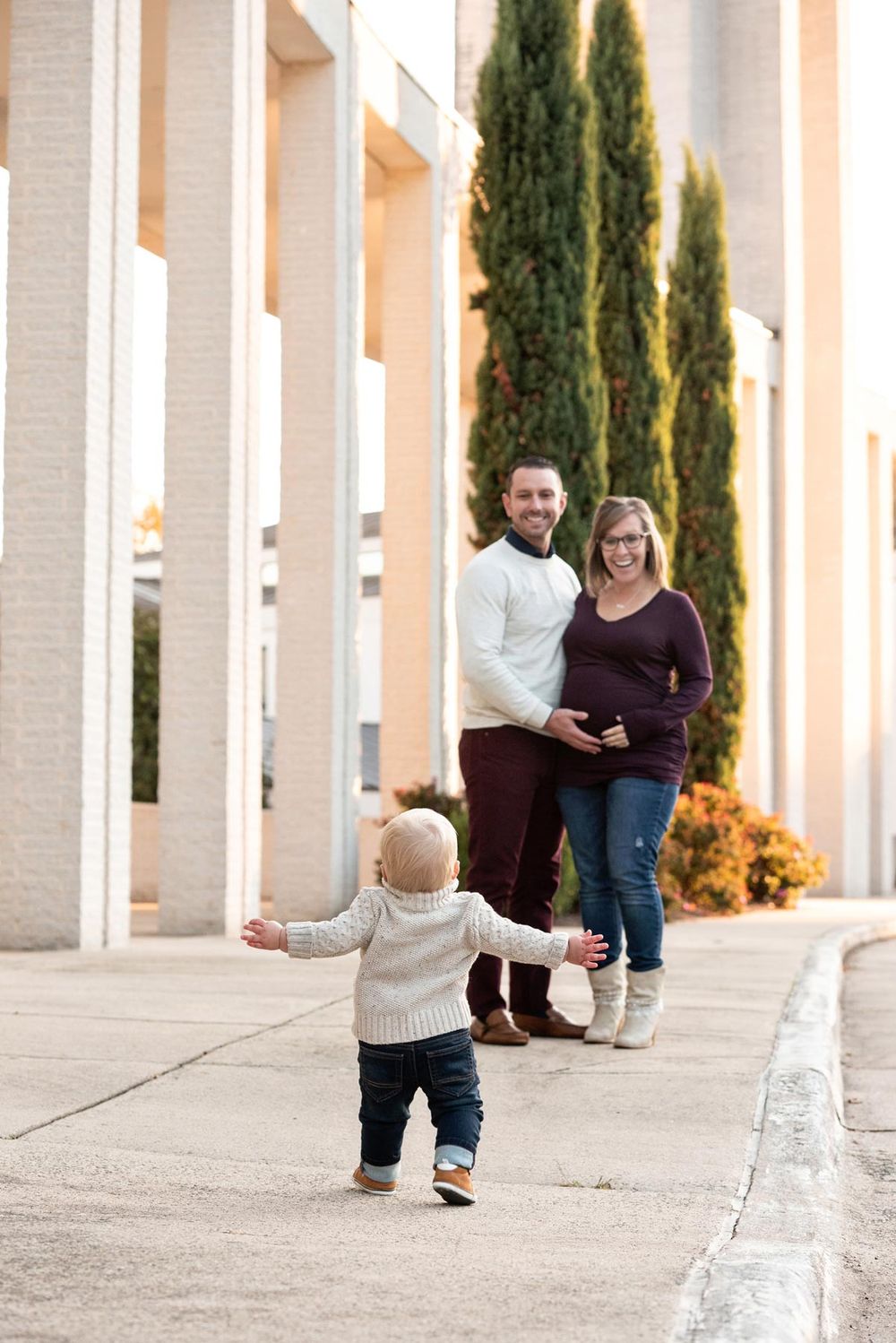 A small toddler in a sweater walks confidently toward a couple standing by tall cypress trees and white columns.