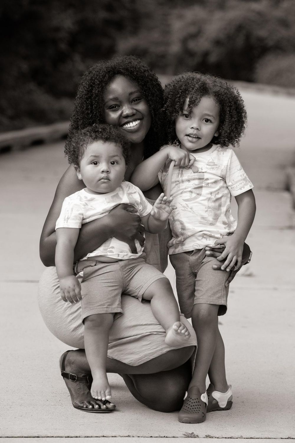 Black and white portrait of a mother kneeling with two small children with curly hair on a path outdoors.