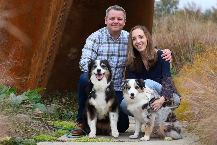 Two Australian Shepherds sitting with their owners in front of a rustic metal structure outdoors.