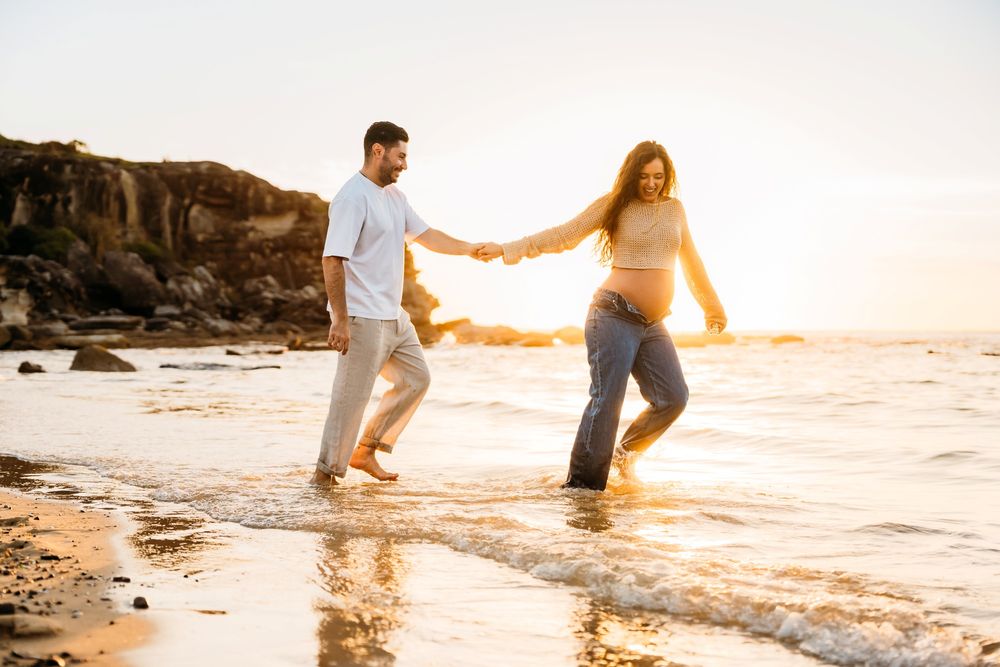 A couple holds hands while walking along a rocky beach at sunset with golden light illuminating the scene for their maternity photoshoot