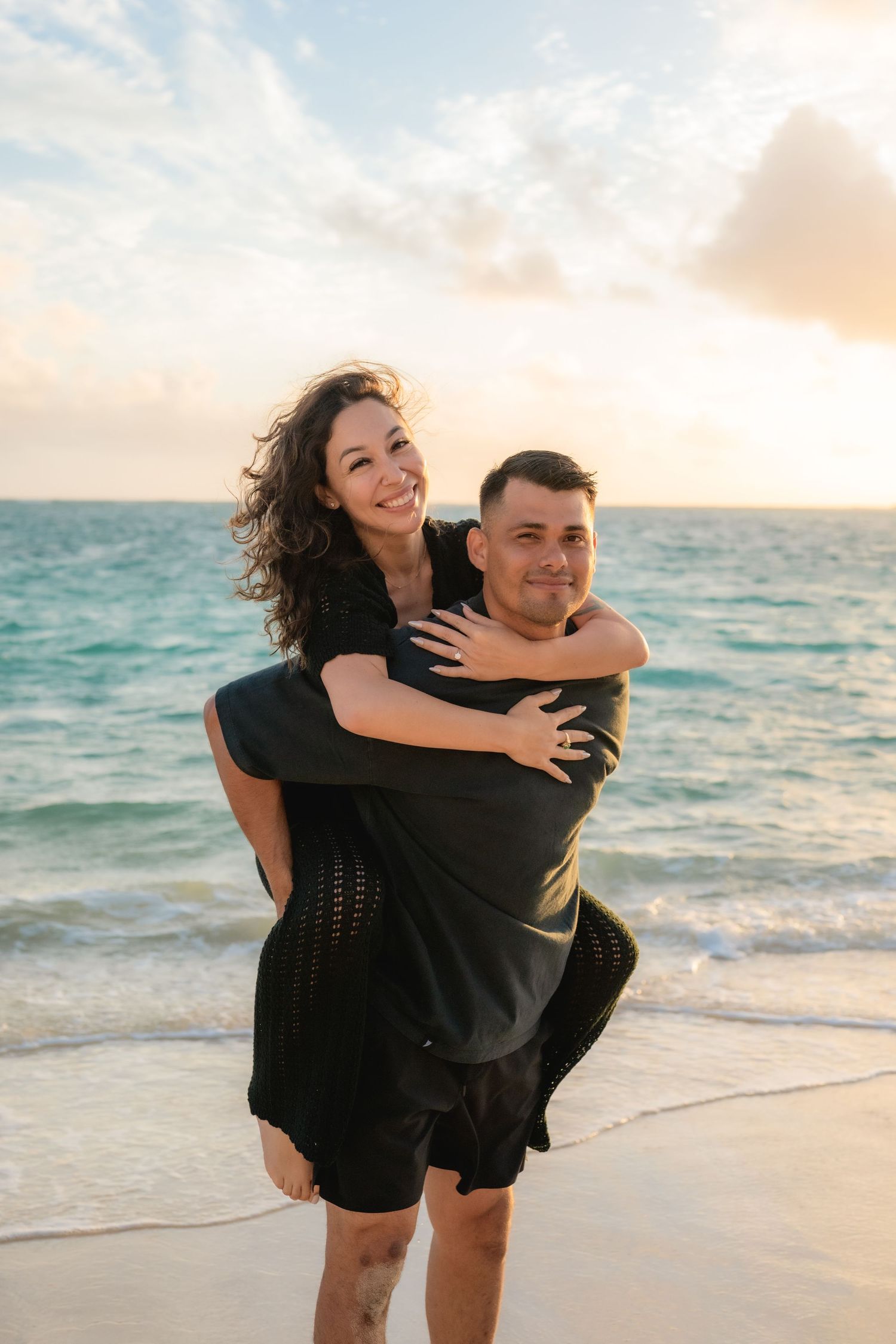 women being carried on man's back with both of them smiling at the camera. on a beach at sunrise.