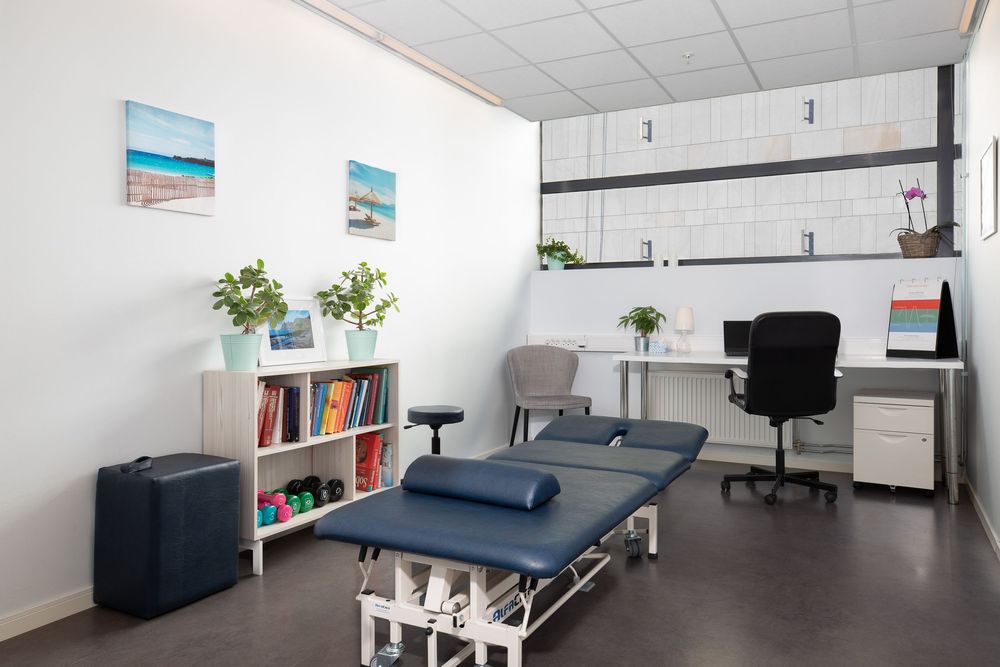 Modern physiotherapy treatment room with examination table, bookshelf, desk, and coastal artwork on white walls.