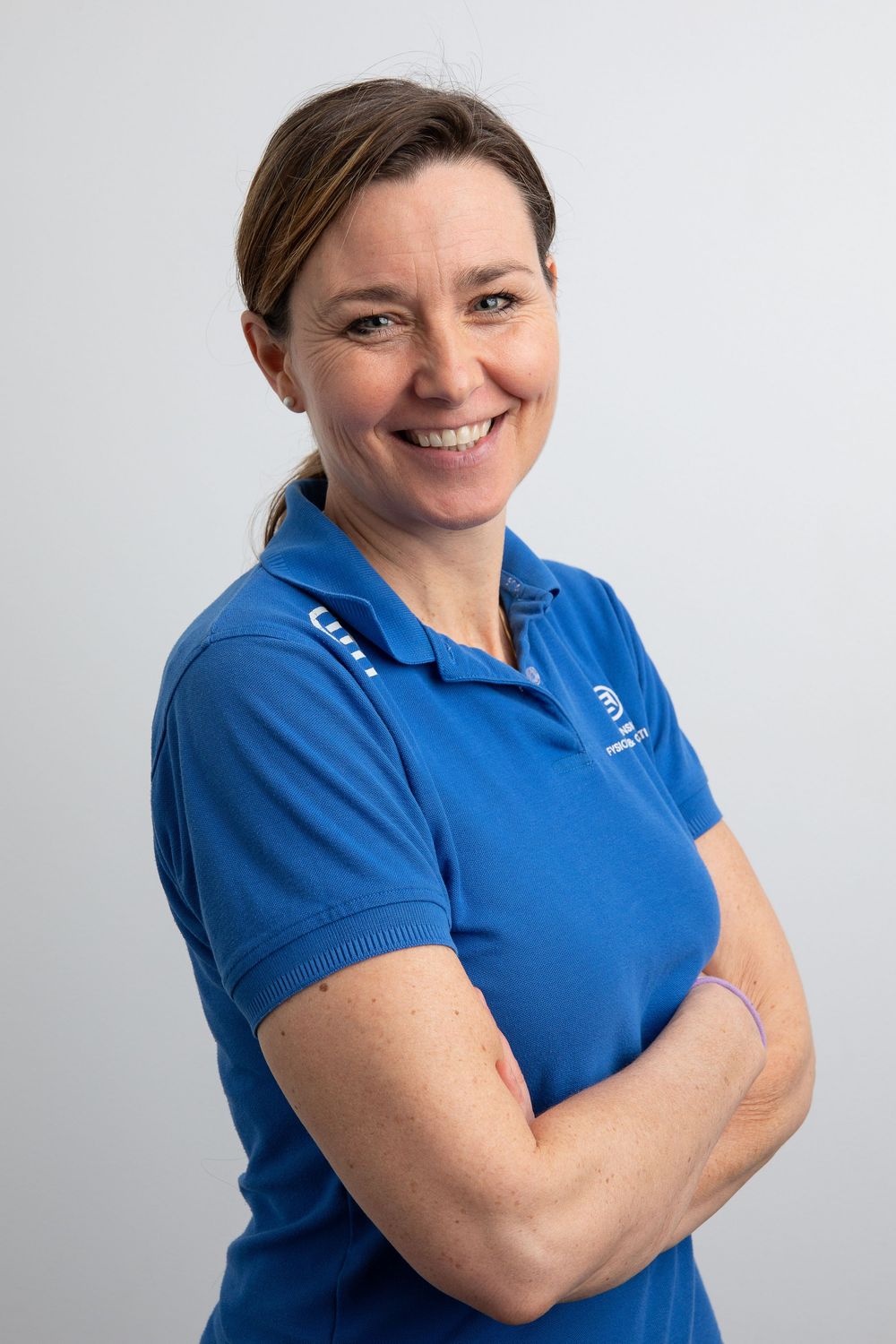 Smiling woman in blue polo shirt with arms crossed poses confidently against a light gray background.