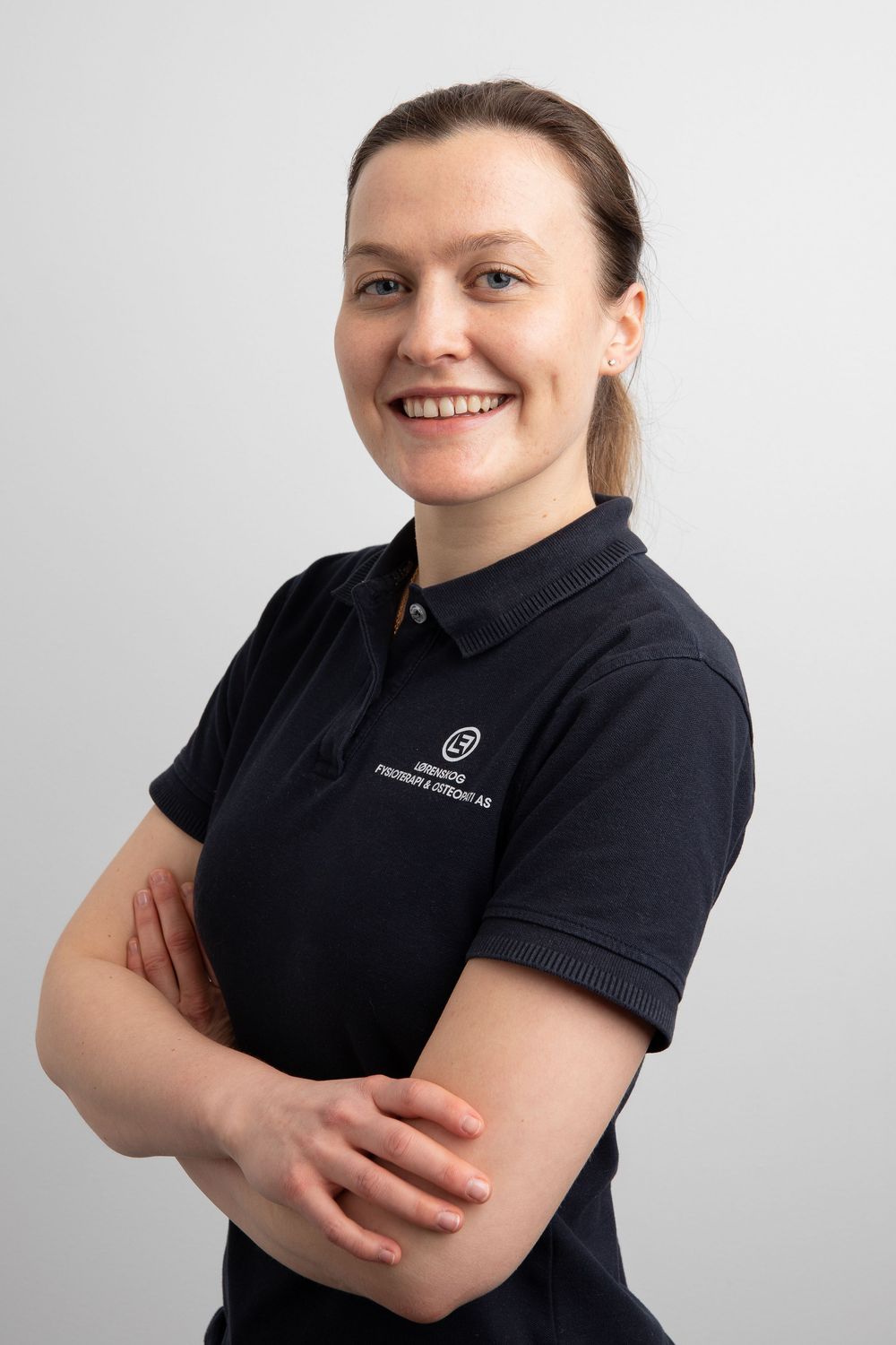 Smiling female physiotherapist in navy polo shirt with arms crossed against light gray background.