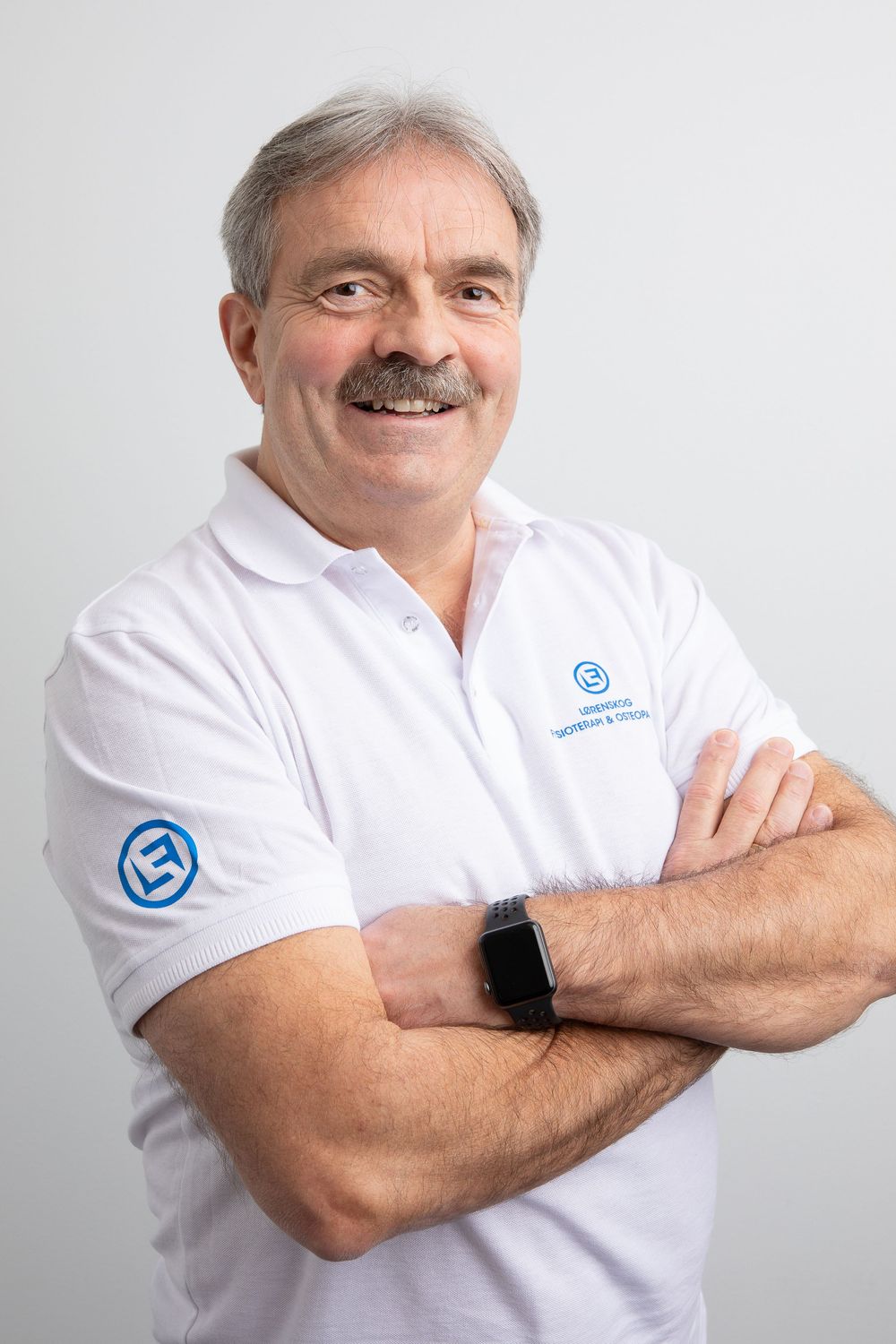 Smiling middle-aged man with mustache, arms crossed, wearing white polo shirt with blue logo and smartwatch.