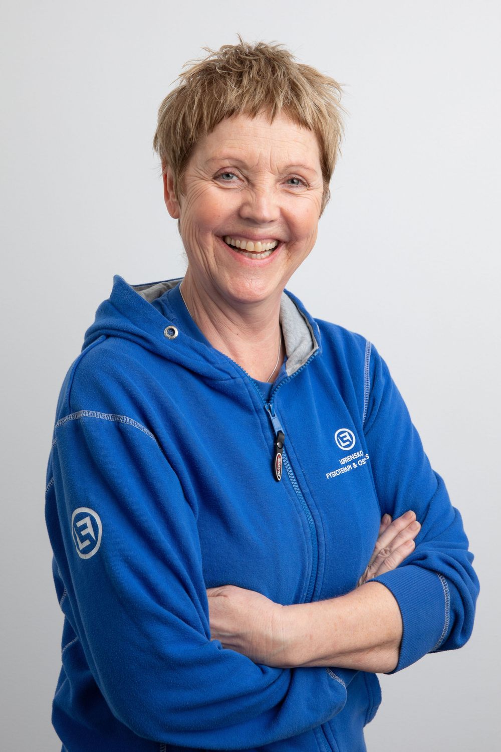 Smiling woman in blue zip-up hoodie with crossed arms, short blonde hair, professional portrait.