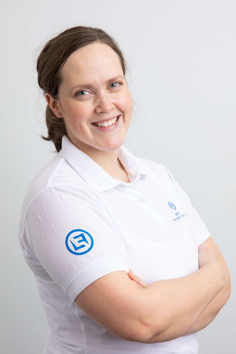 Smiling woman in white polo shirt with blue logo, arms crossed, professional headshot on light gray background.