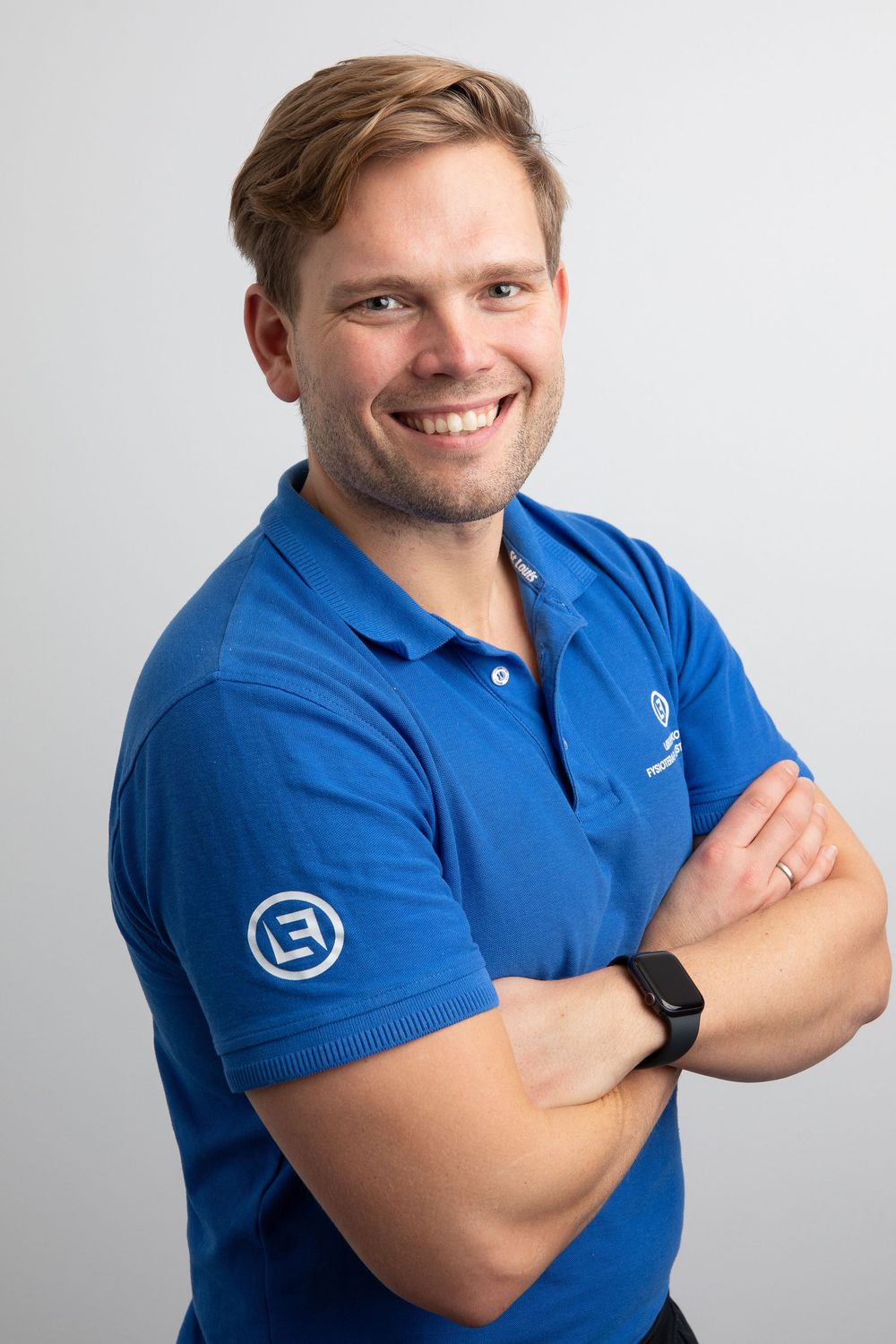 Smiling young man in blue polo shirt with logo, arms crossed, wearing smartwatch, professional headshot on gray background.