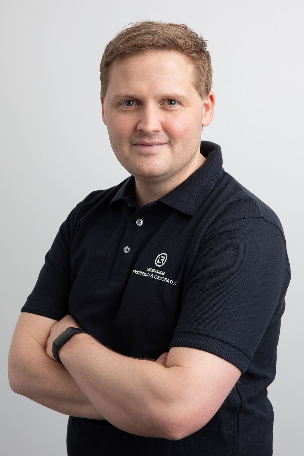 Professional man in black polo shirt with arms crossed, smiling confidently against a light gray background.