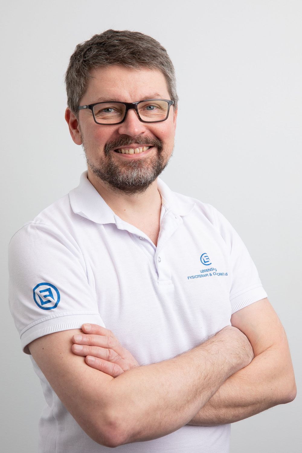 Smiling man with glasses and beard, arms crossed, wearing a white polo shirt with a company logo.