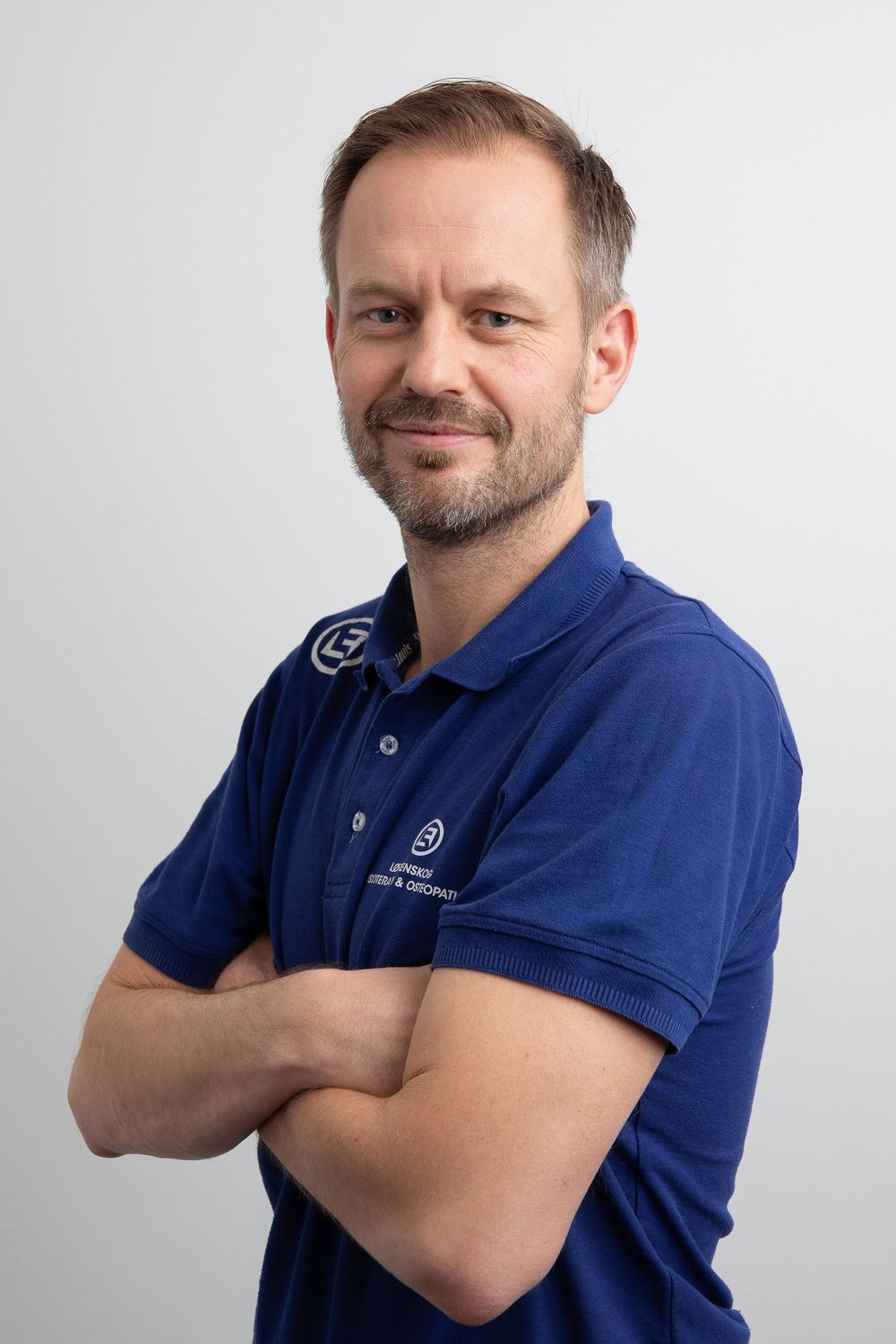 Professional man in blue polo shirt with arms crossed, smiling confidently against a light gray background.
