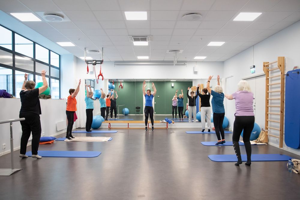Group fitness class with participants doing yoga stretches on blue mats in a bright modern studio with mirrored walls.
