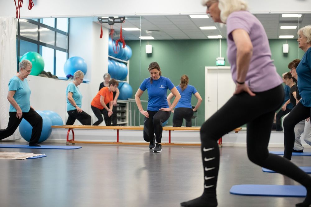 Group fitness class with instructor leading exercises using stability balls in a bright gym with mirrored walls.