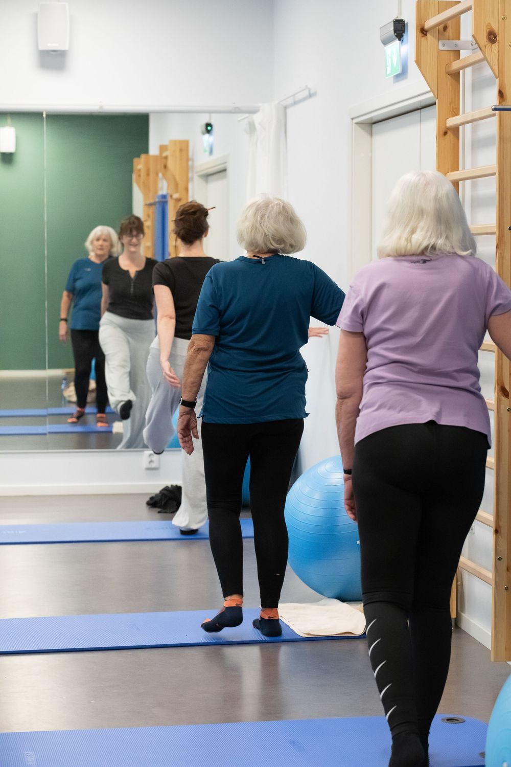 Senior women exercising in a fitness class, walking on blue mats in a bright gym with mirrors and balance equipment.