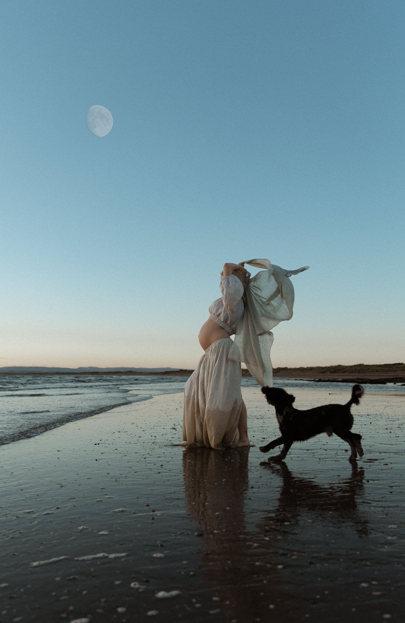 Pregnant woman standing barefoot on wet sand at the shoreline beneath a pale moon, with her dog beside her.