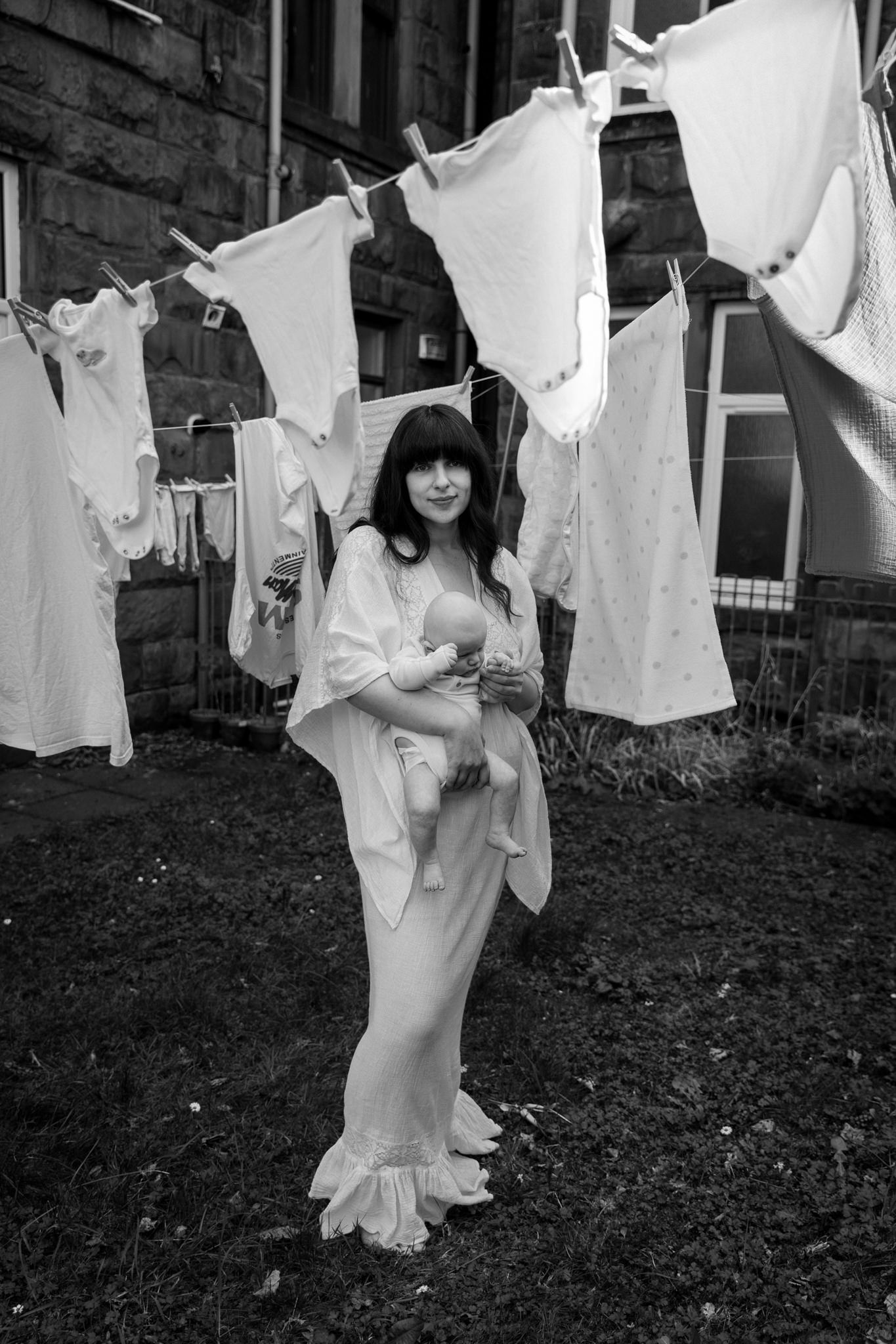 Mother holding her baby beneath hanging laundry in front of Glasgow tenement buildings, wearing a white lace dress.