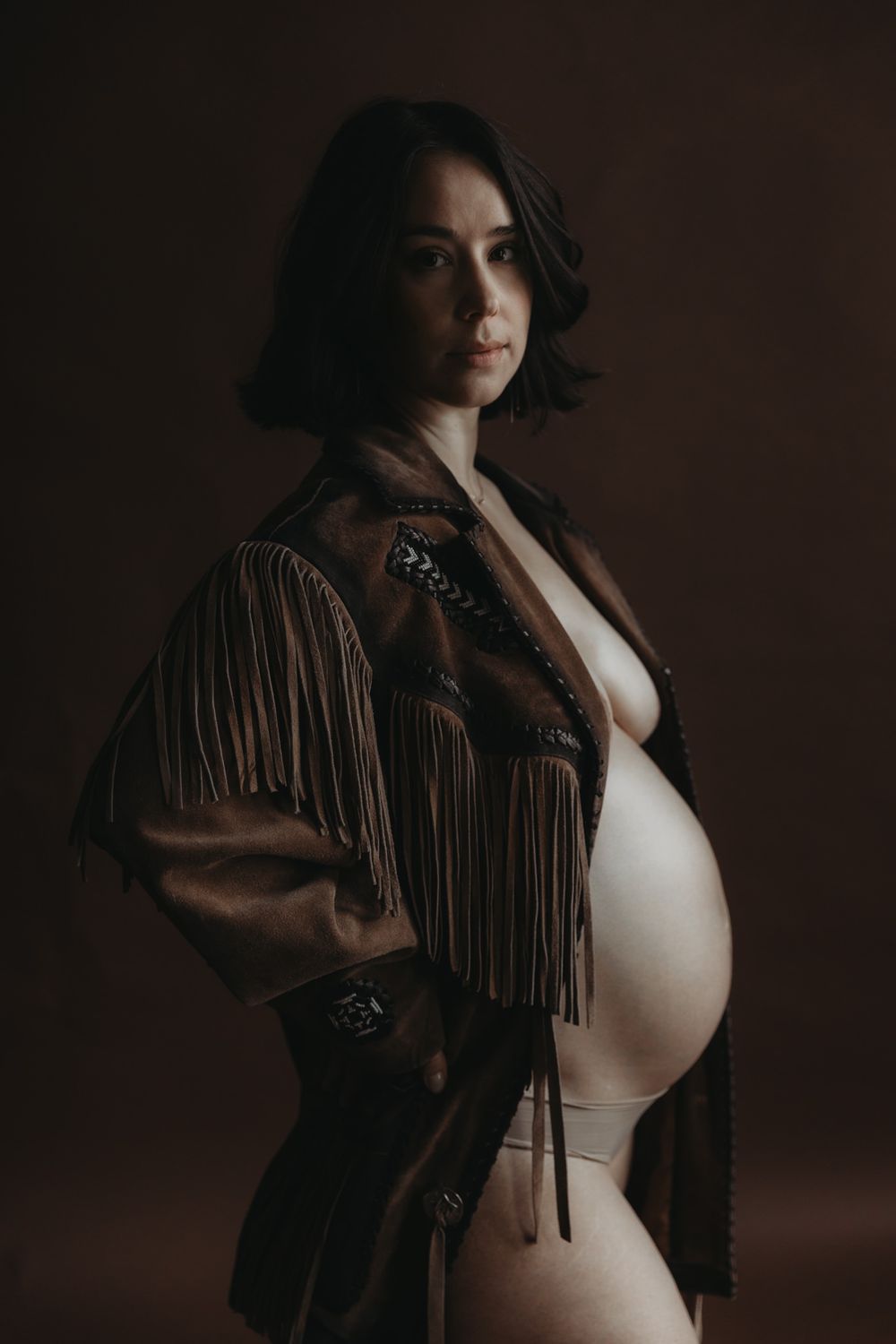 Pregnant woman photographed in a studio setting against a dark background, wearing a fringed jacket