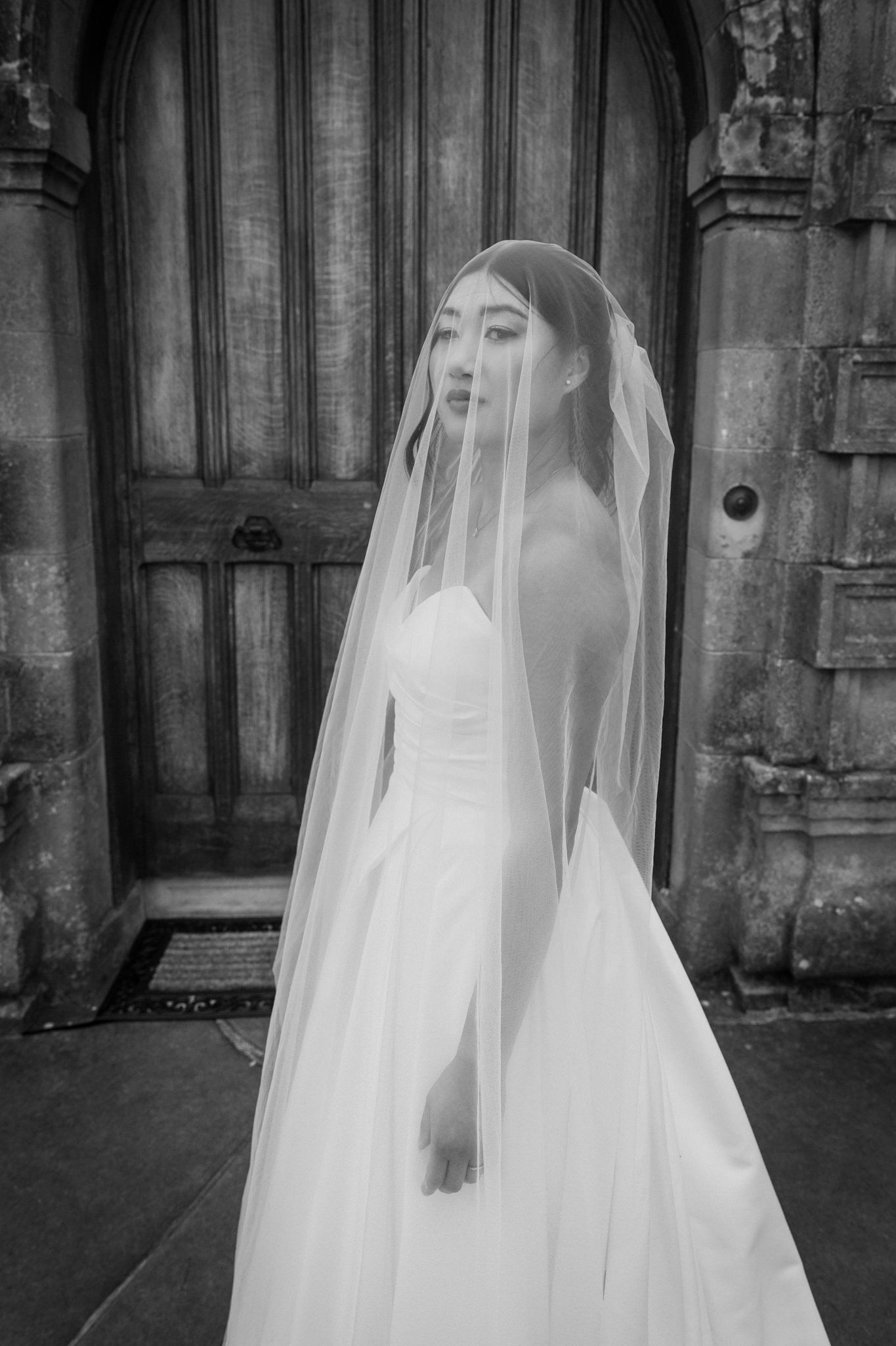 Bride wearing a veil standing in front of a stone doorway, photographed in black and white