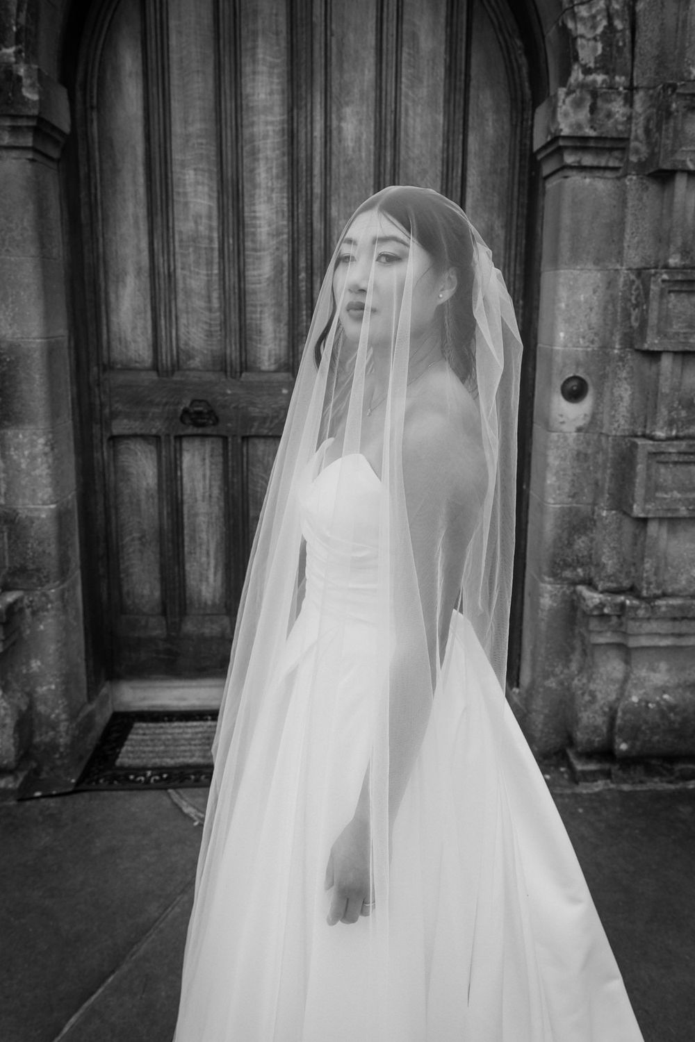 Bride wearing a veil standing in front of a stone doorway, photographed in black and white