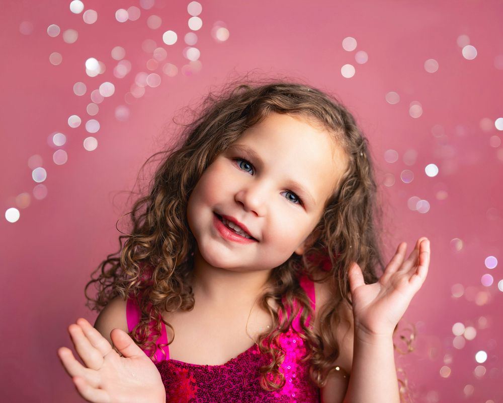 A joyful young girl with curly hair in a pink sequin dress poses against a pink bokeh background.