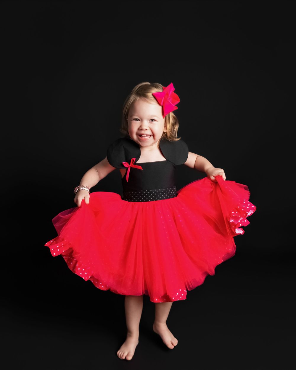 Smiling toddler girl in red tutu dress with black bodice and pink bow hair clip poses barefoot.