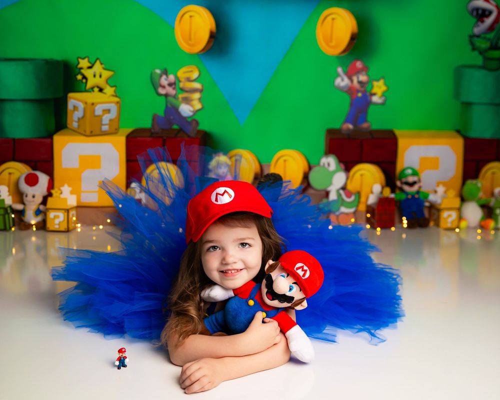 Girl in Mario costume with tutu holds Mario plush doll in front of Super Mario Bros themed backdrop.