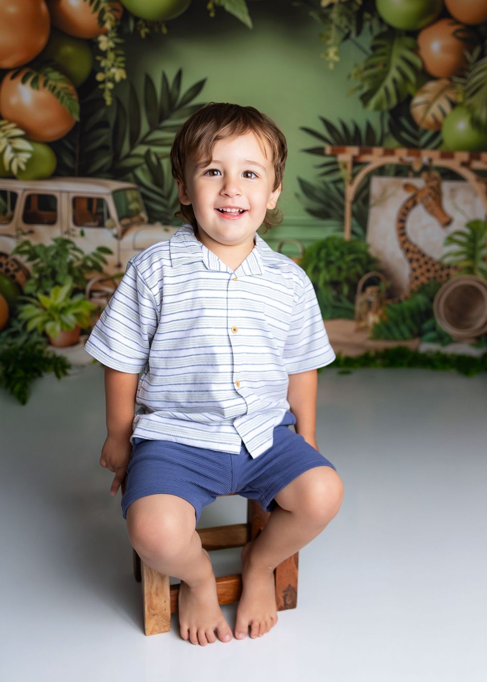 Young child wearing striped white shirt and blue shorts sits barefoot on wooden stool against tropical backdrop.