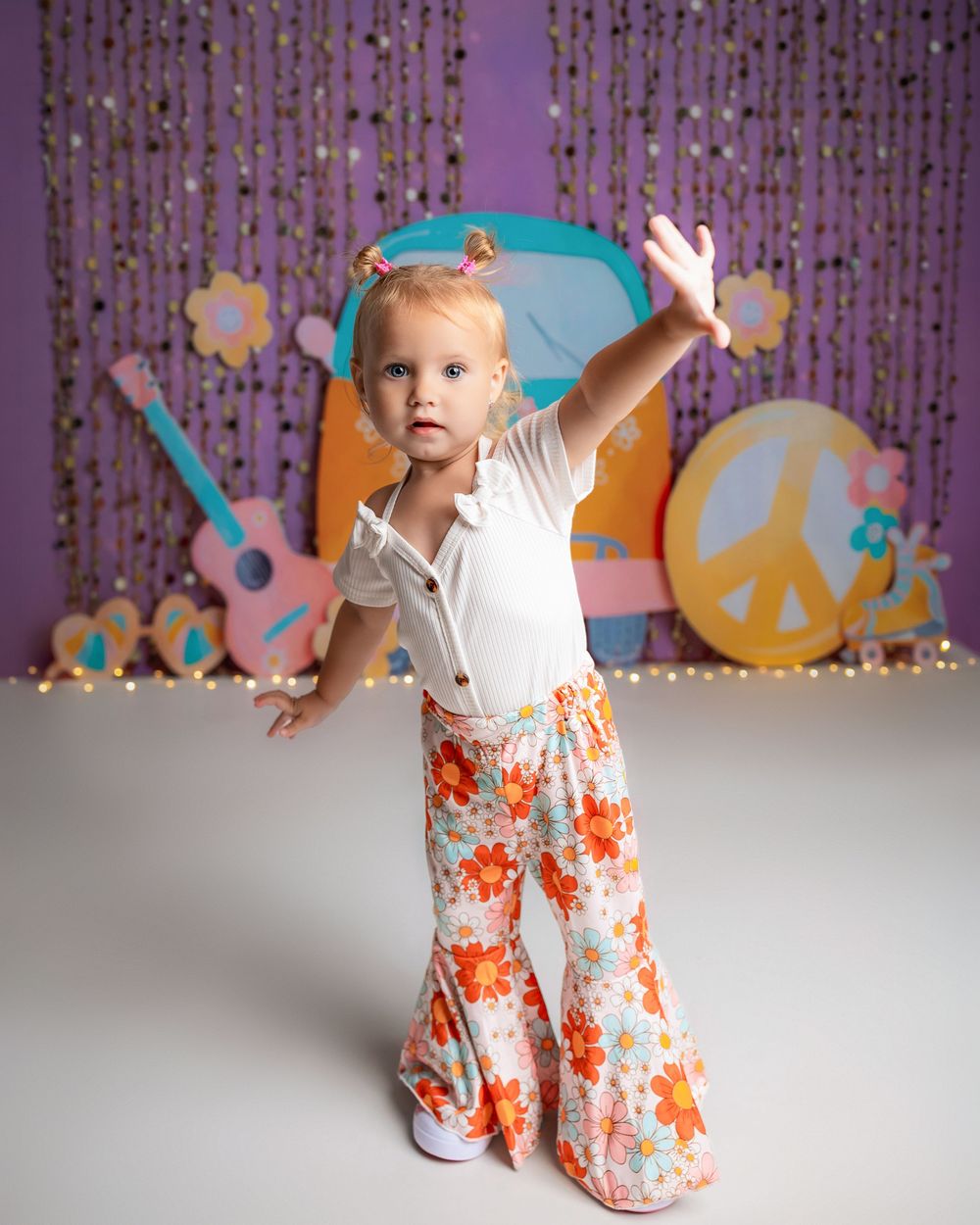 Toddler girl in floral bell-bottom pants and white top poses with arms raised before a colorful retro 70s-themed backdrop.