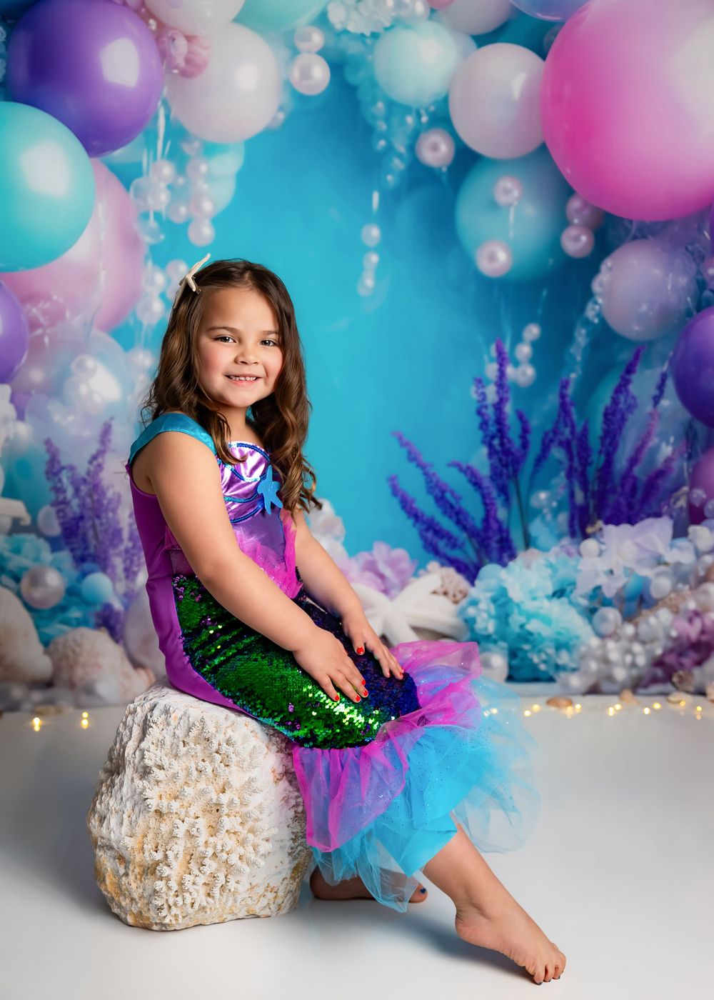 Young girl in mermaid costume sitting on rock with colorful balloon arch and underwater themed backdrop.