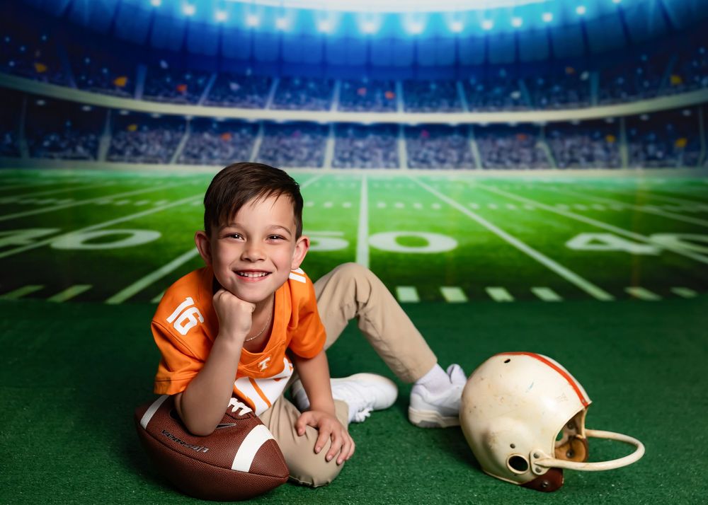 Young boy in orange football jersey poses with football and helmet on green field backdrop in stadium setting.