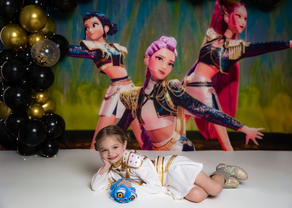 Toddler girl in white outfit poses at birthday party with colorful cartoon character backdrop and black gold balloons.