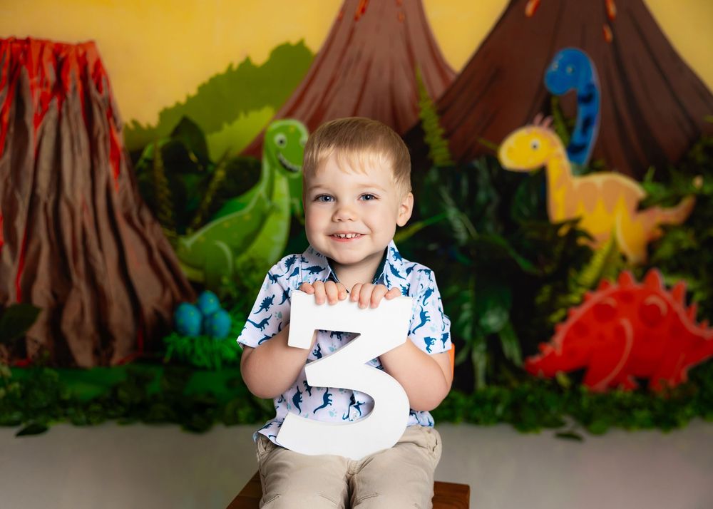 A child poses with colorful dinosaur decorations and volcanoes in a themed photo backdrop setting.