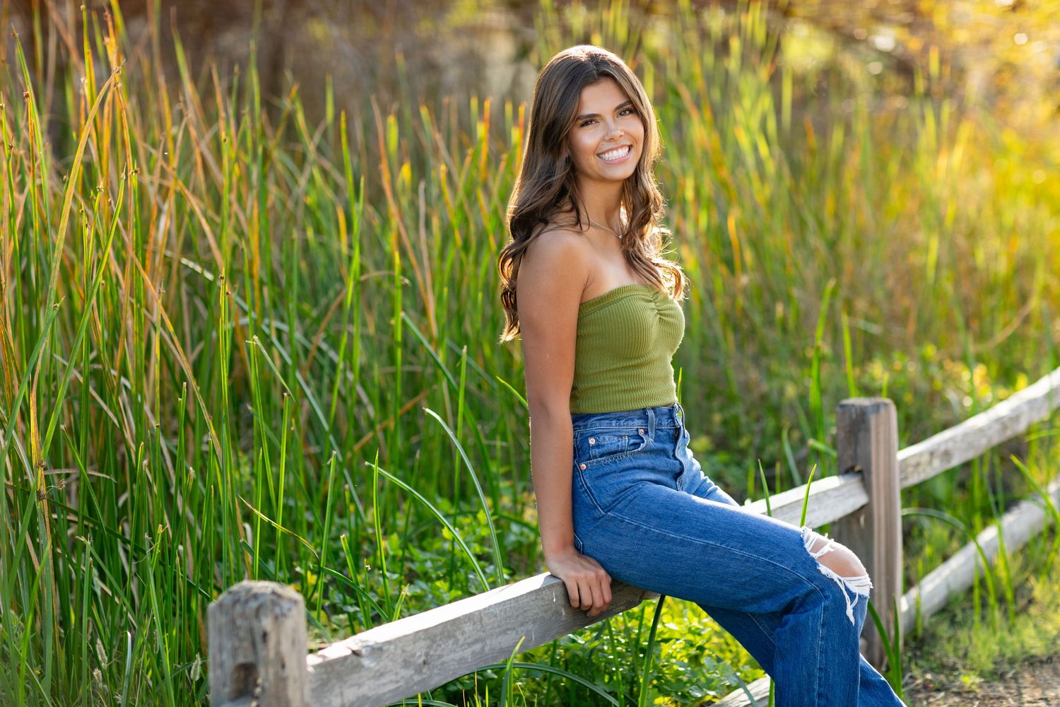A high school senior sits on a wooden fence at sunset wearing a green top and blue jeans with tall grass in the background.