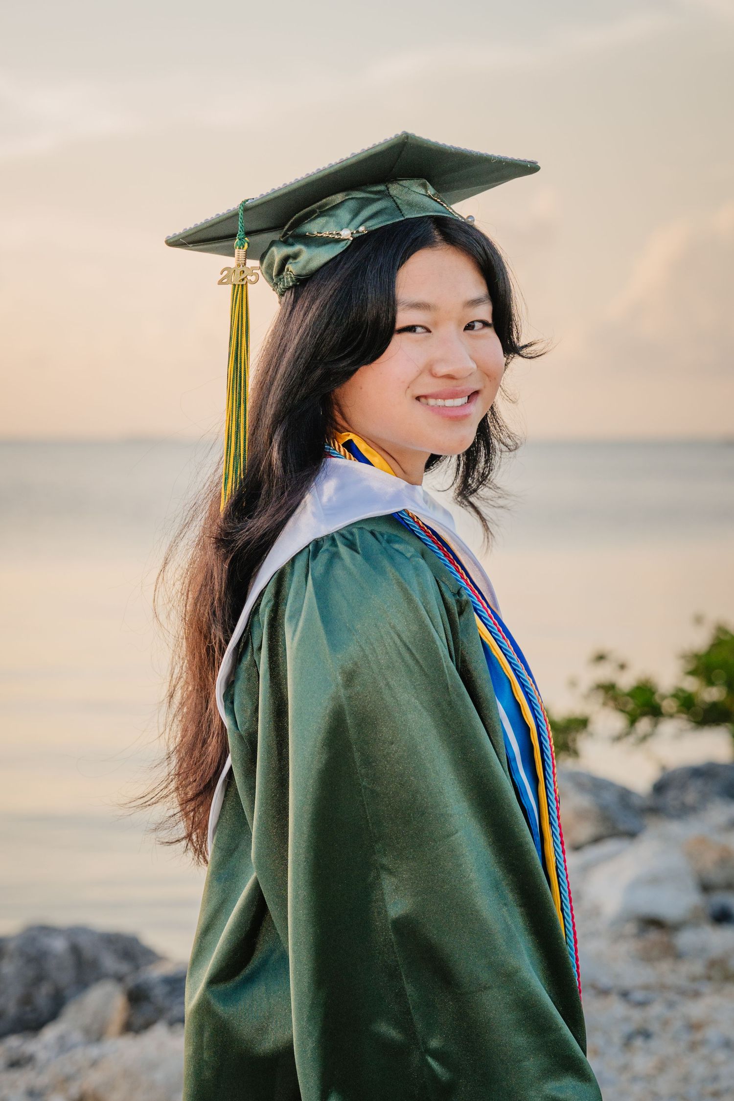 A graduate in a green cap and gown smiles while posing for photos at sunset by a rocky coastline.