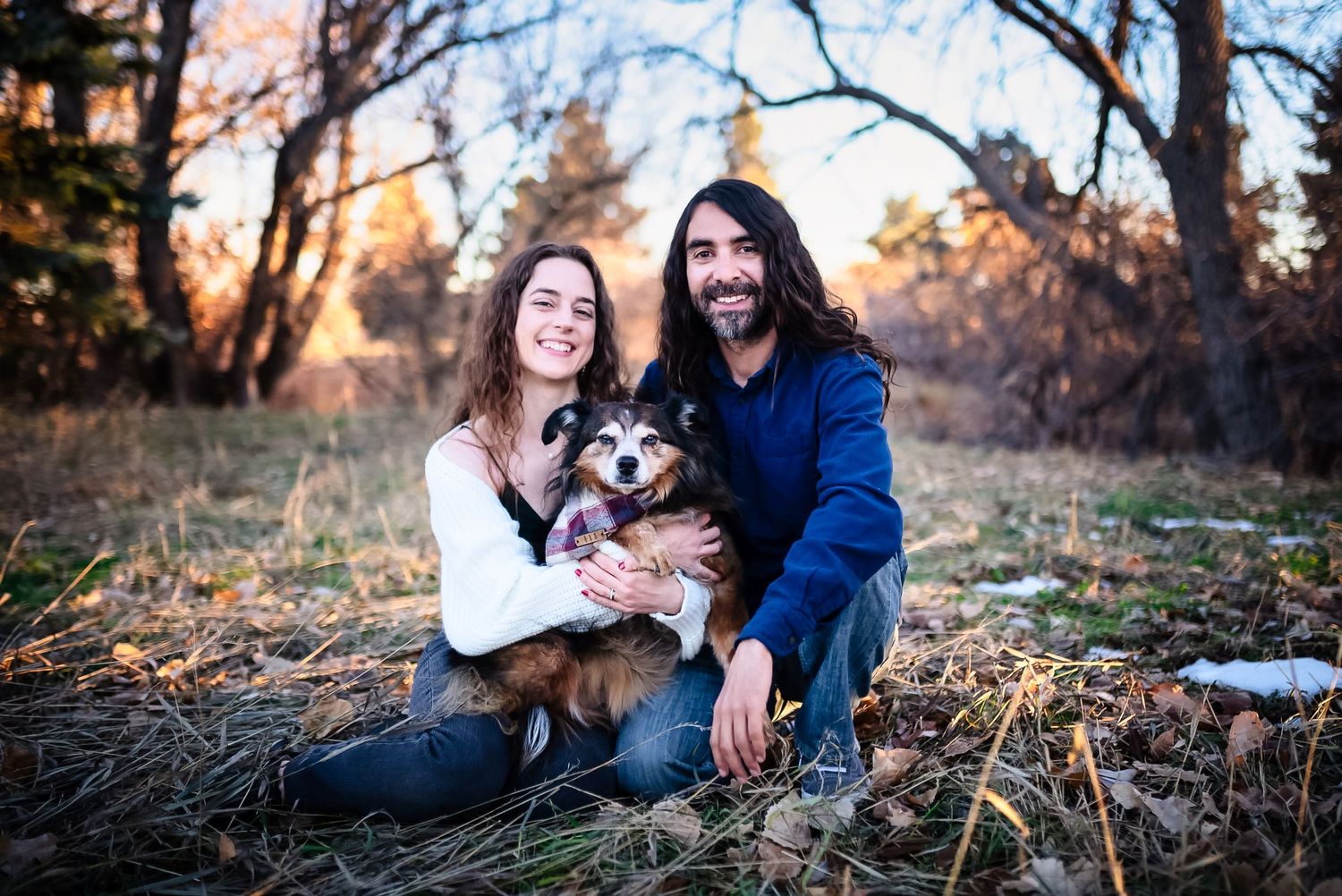 A couple sits outdoors with their mixed breed dog on an autumn day during golden hour.