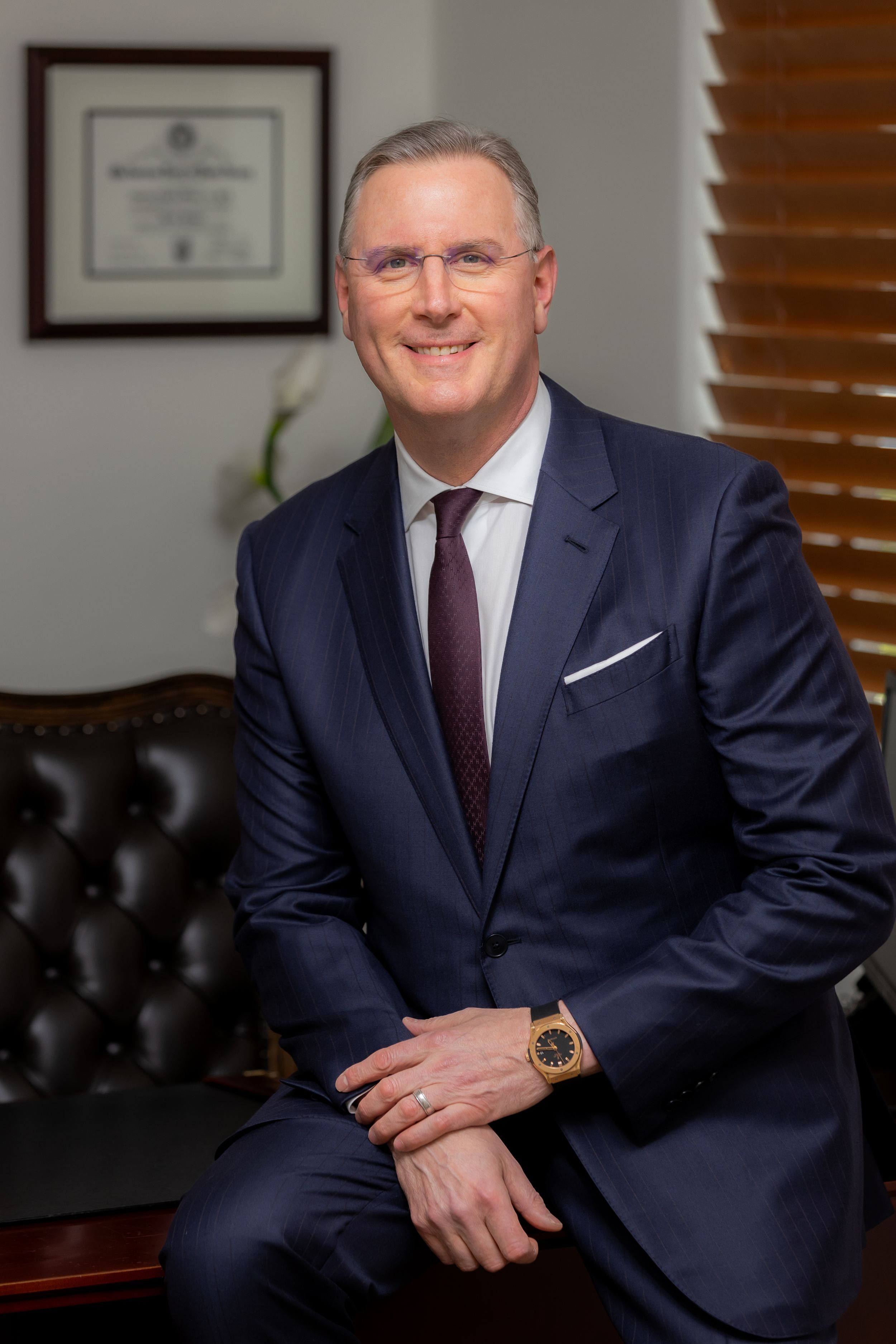 Professional business portrait in navy suit with burgundy tie seated in front of office wall with framed certificates.