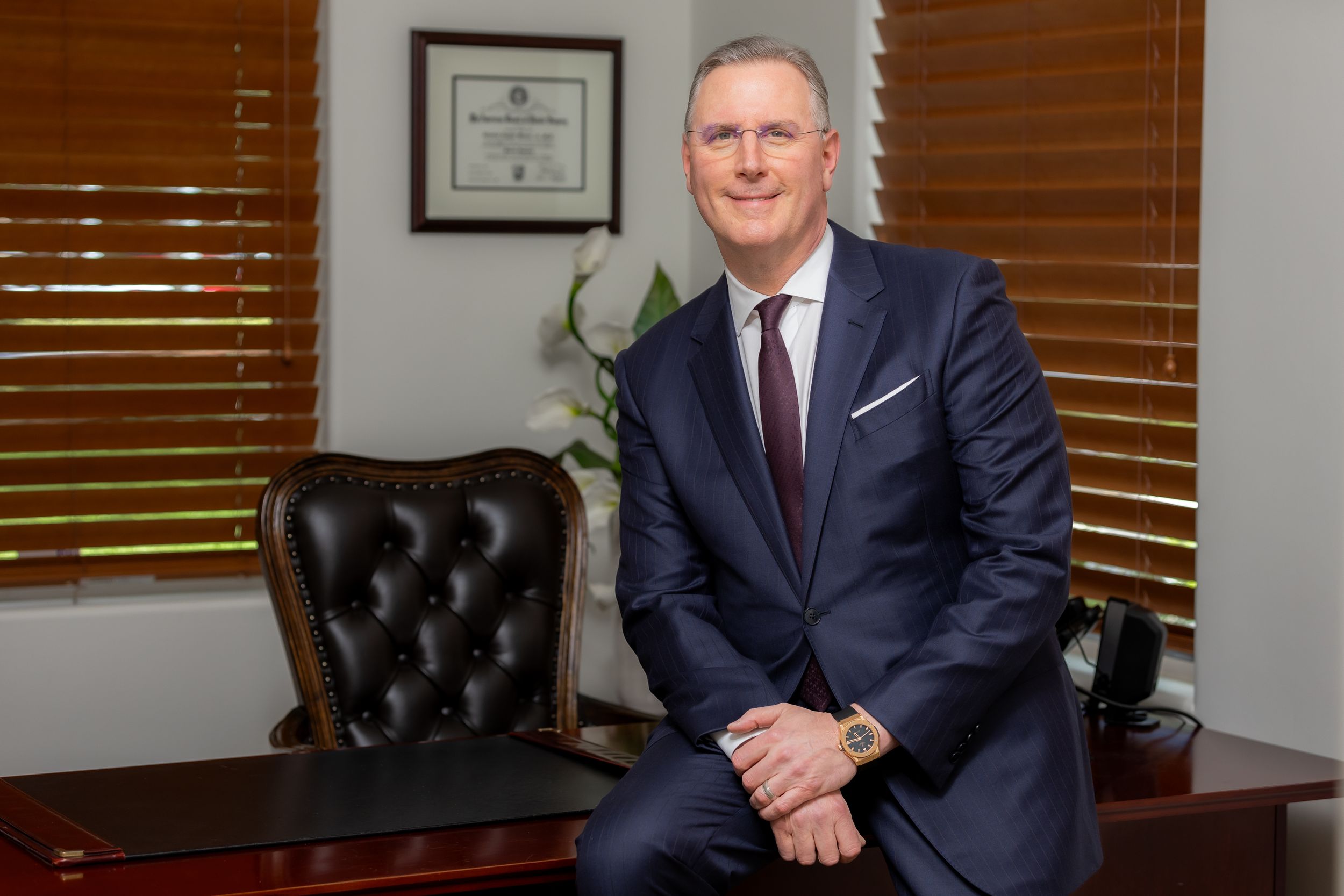 Professional executive seated at desk wearing navy suit and burgundy tie in office with wooden blinds.
