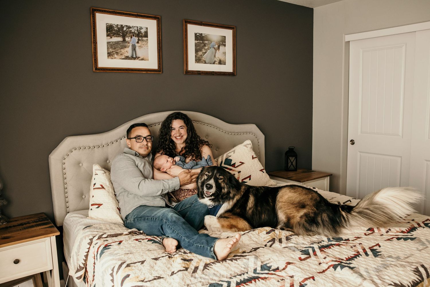 Relaxed family portrait on bed with dogs and baby in cozy bedroom setting.