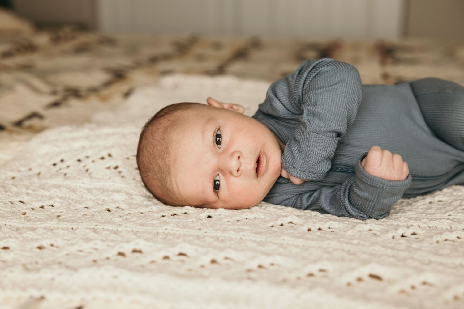Baby in grey outfit lying on white textured bedding looking at camera with bright eyes.