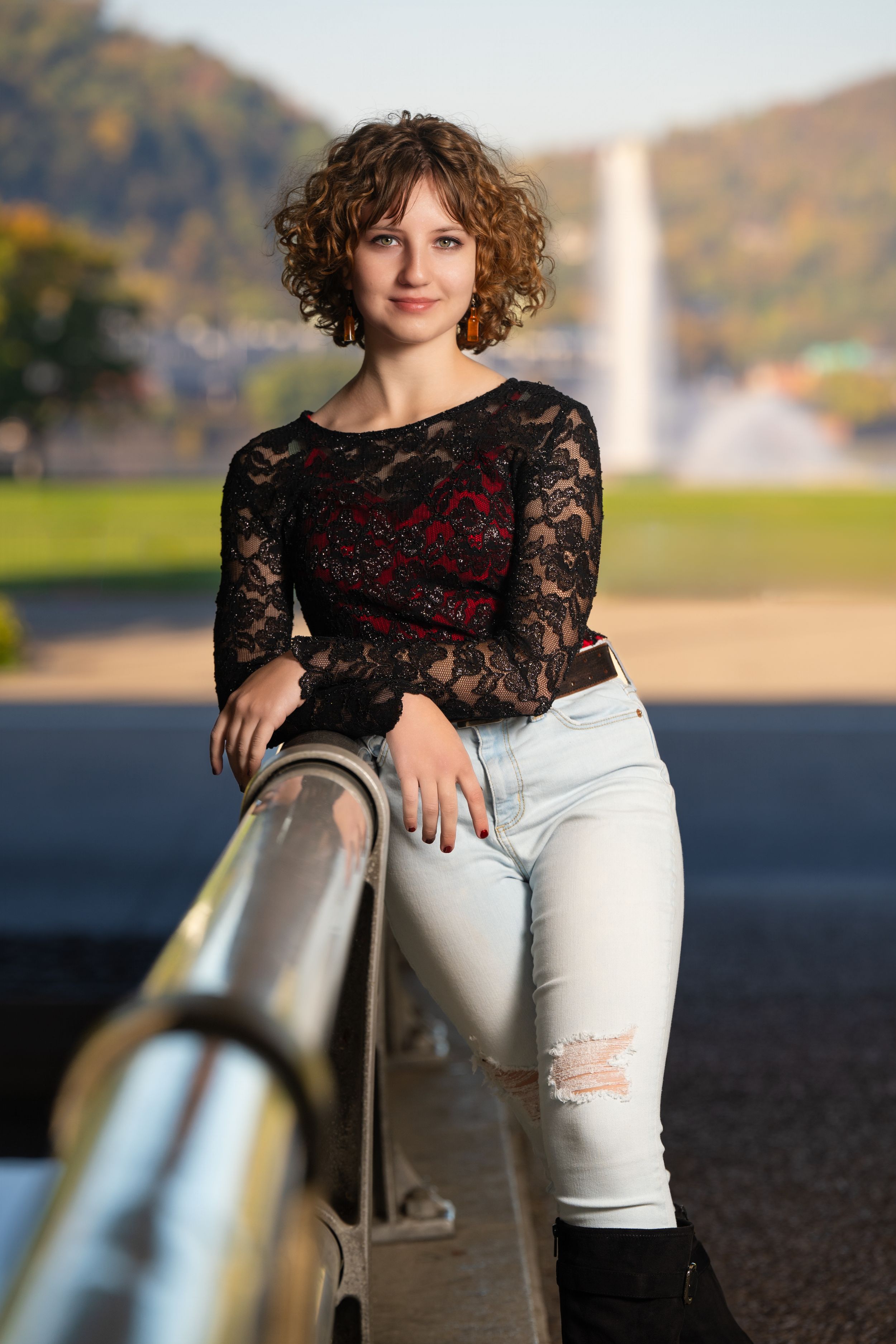Person in black lace top and white jeans poses casually against metal railing during outdoor autumn photoshoot.