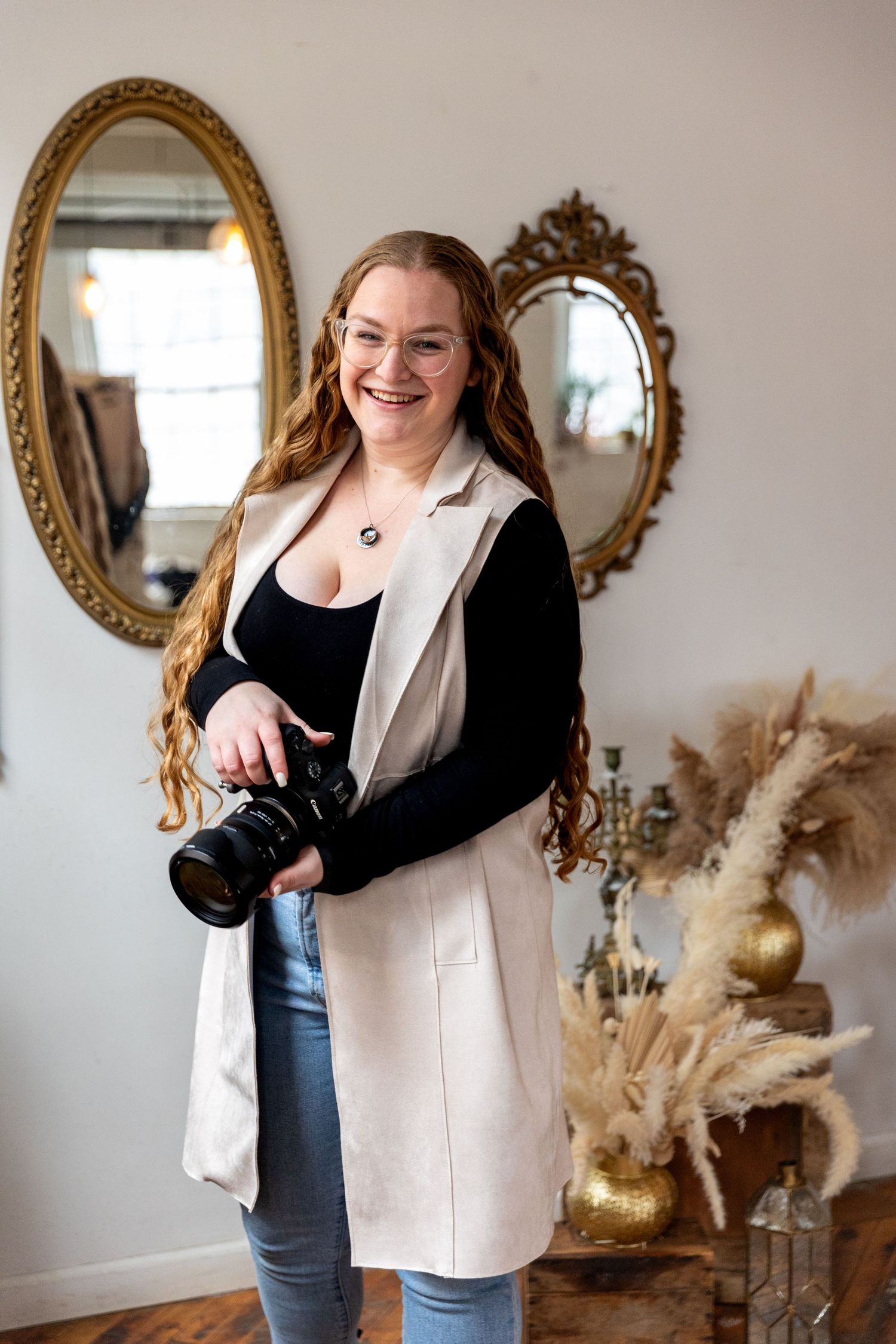 Smiling female photographer with long red hair holding a camera in a boho-styled studio with ornate mirrors.