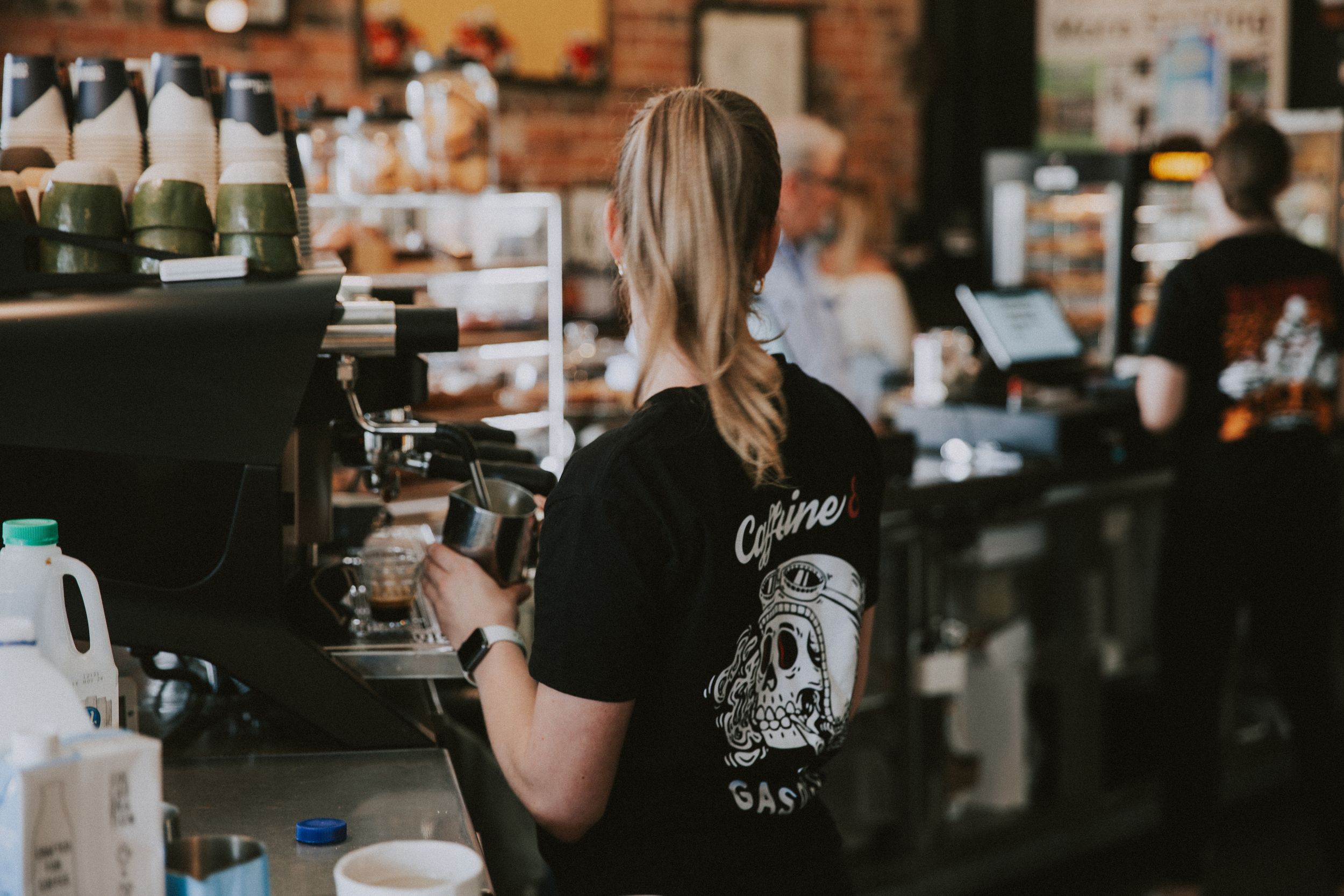A coffee shop barista wearing black prepares drinks behind a wooden counter in a busy cafe setting.