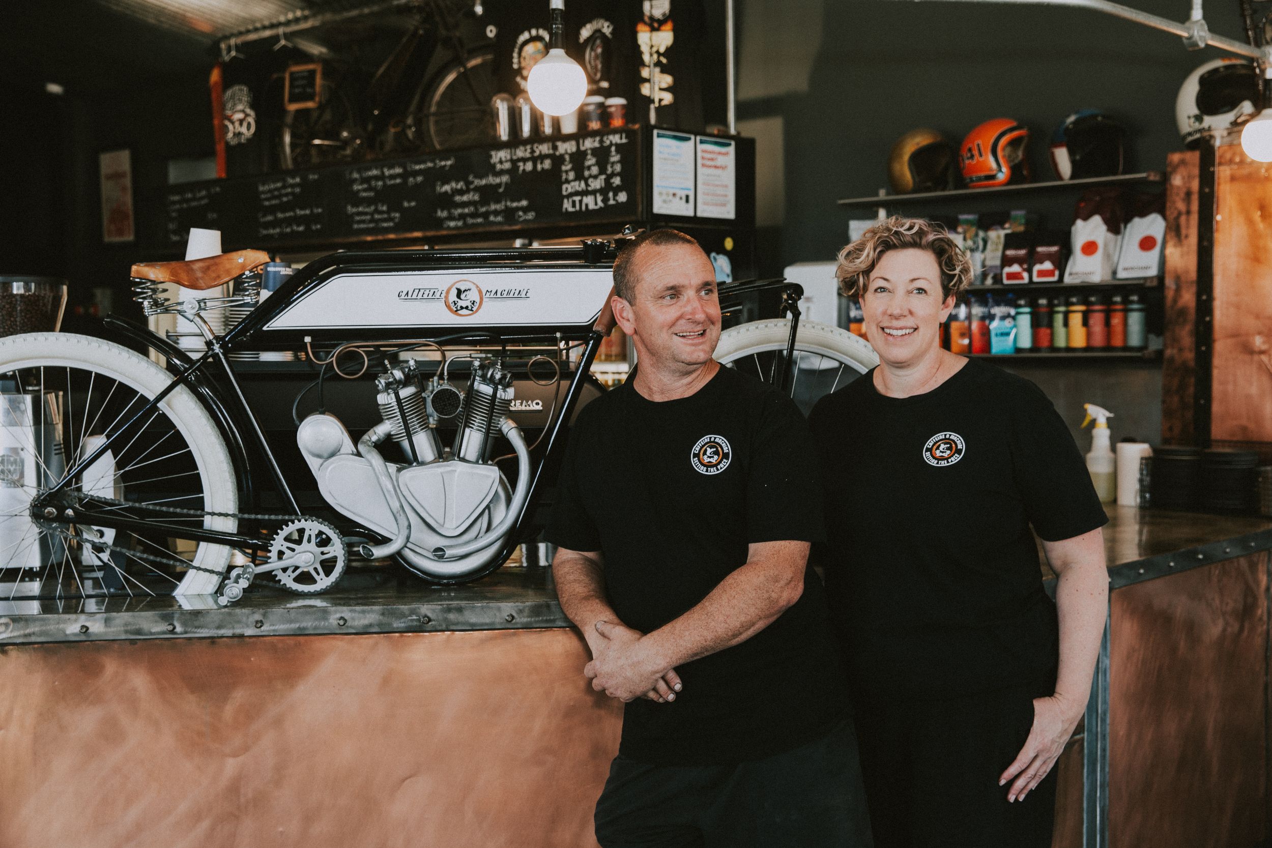 Two staff members in black t-shirts stand in front of a vintage motorcycle display at a shop.
