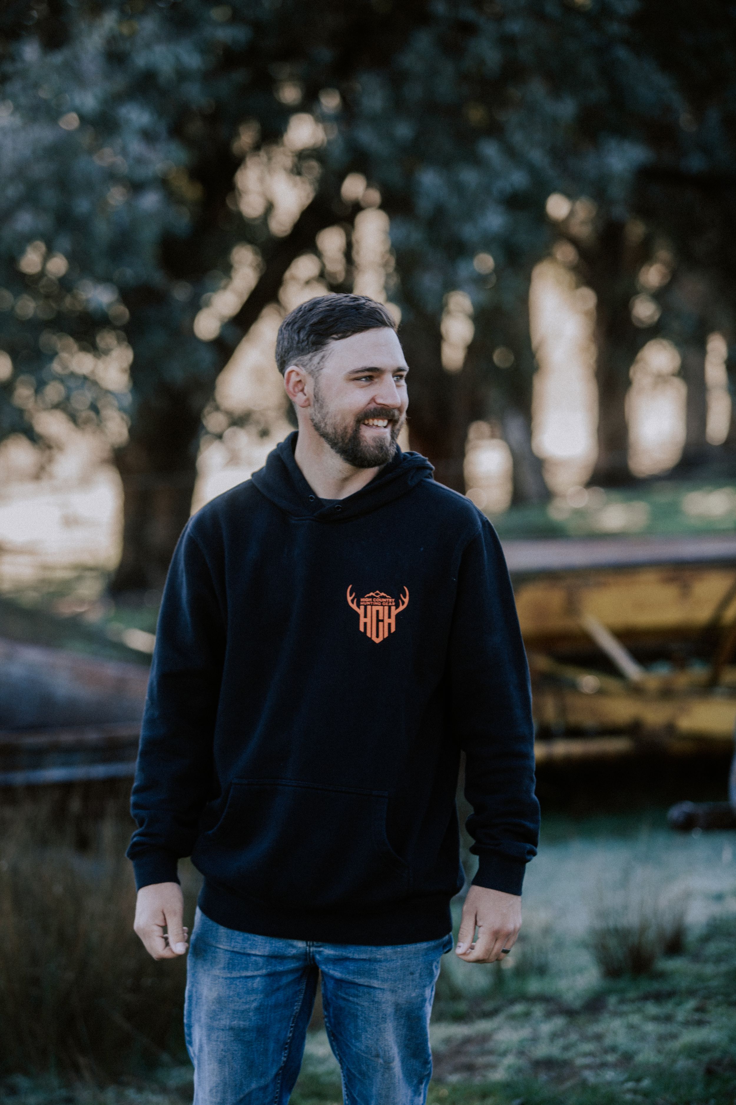 A person in a black sweatshirt and jeans stands in a park setting during golden hour lighting.
